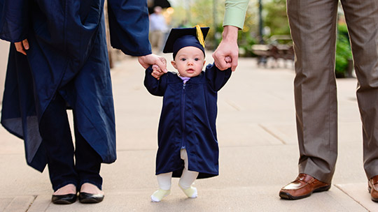 Baby in cap and gown holding parents’ hands