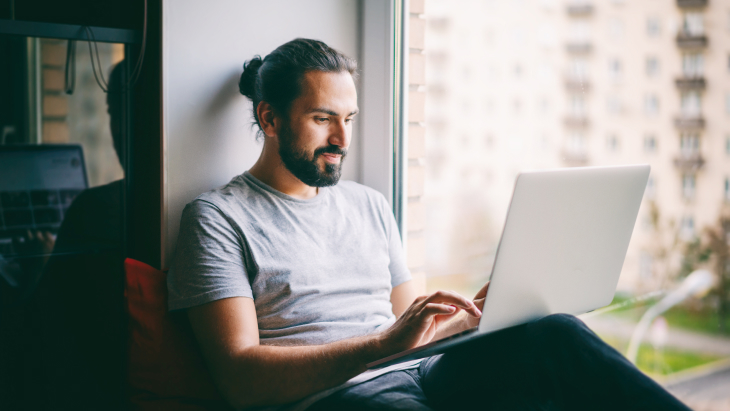 Life Insurance for Freelance Hero Desktop | Handsome attractive young dark-haired man working on laptop at home while sitting on windowsill with city view