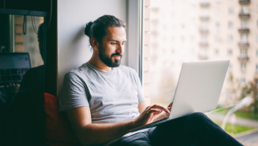 Life Insurance for Freelance Hero Mobile | Handsome attractive young dark-haired man working on laptop at home while sitting on windowsill with city view