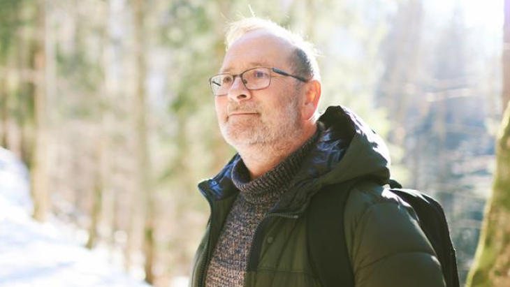 Outdoor portrait of middle age 55 - 60 year old man hiking in winter forest, wearing warm jacket and black backpack