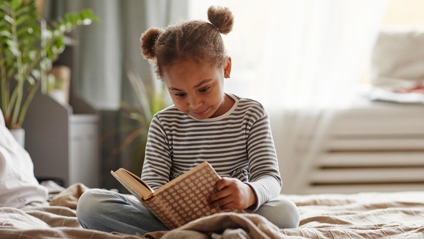 Portrait of cute young girl reading book while sitting on bed in a cozy room.