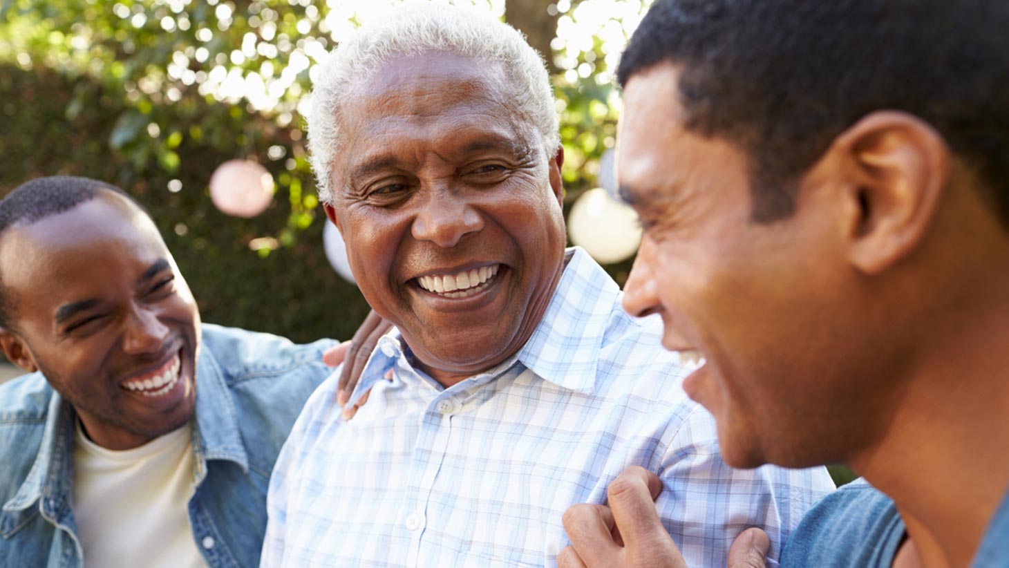 Older man sitting in garden with his two adult sons smiling and chatting during a nice day.