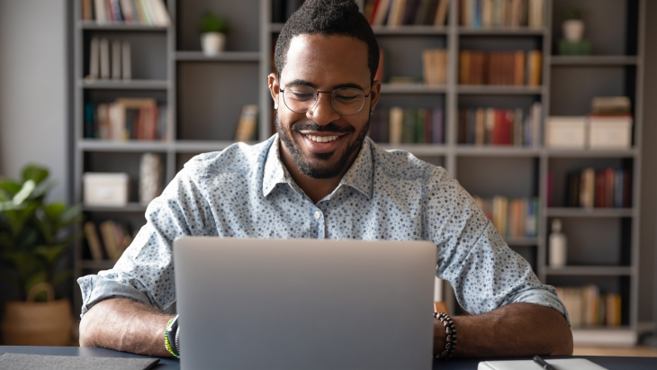 Front view head shot young smiling african american businessman in eyewear looking at laptop screen, working on computer sitting at table in office home.